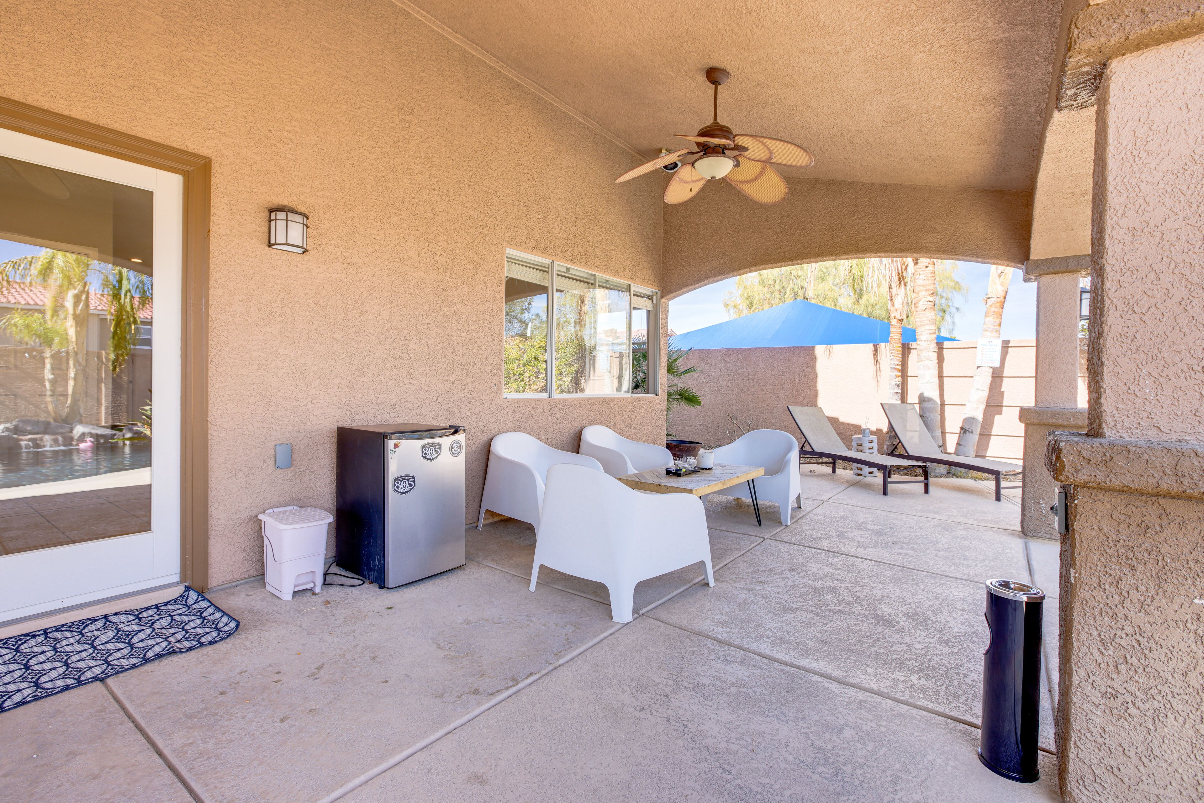 A covered patio with white chairs, a table, a ceiling fan, and a small refrigerator.