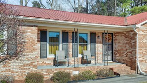 A single-story brick house with a red metal roof, a front porch with two chairs, and a small garden area. - Greenville Home: 2 Mi to Downtown! 1 Mi to Ecu! (Greenville)