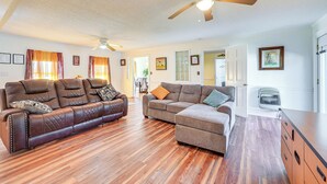 A spacious living room with a brown leather sofa, a gray sectional sofa, and a wooden floor. There are ceiling fans, a door leading to another room, and a wall-mounted air conditioner. - Greenville Home: 2 Mi to Downtown! 1 Mi to Ecu! (Greenville)