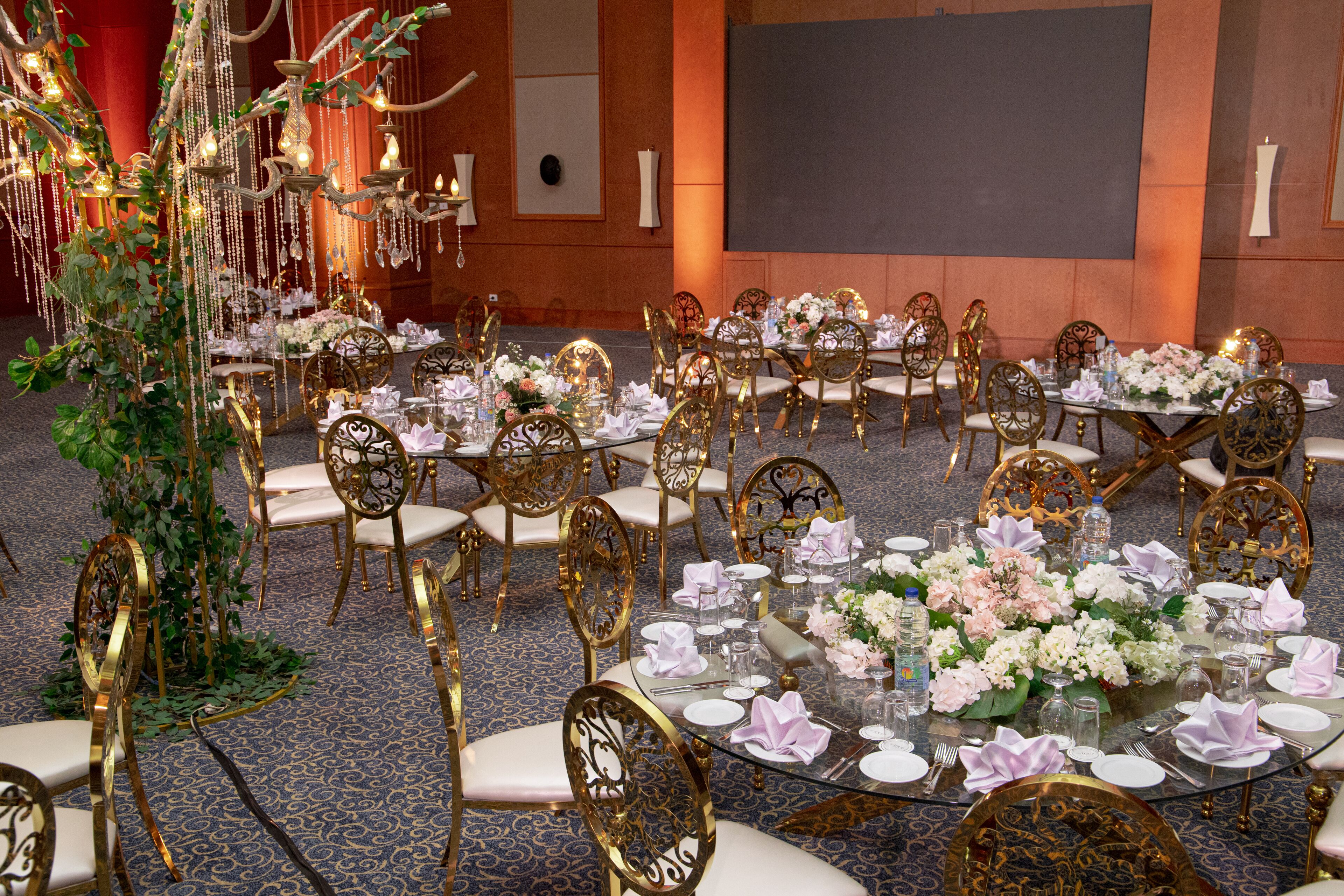 Une salle de banquet avec des tables rondes dressées pour un événement formel, agrément ée de chaises dorées, de nappes blanches et de compositions florales.