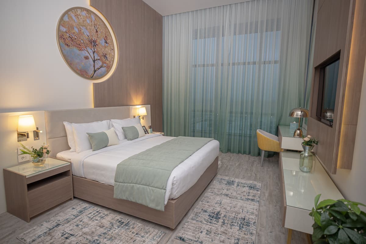 Une chambre d’hôtel moderne, dotée d’un grand lit, de tables de chevet, d’un bureau et d’une grande fenêtre avec des rideaux légers.