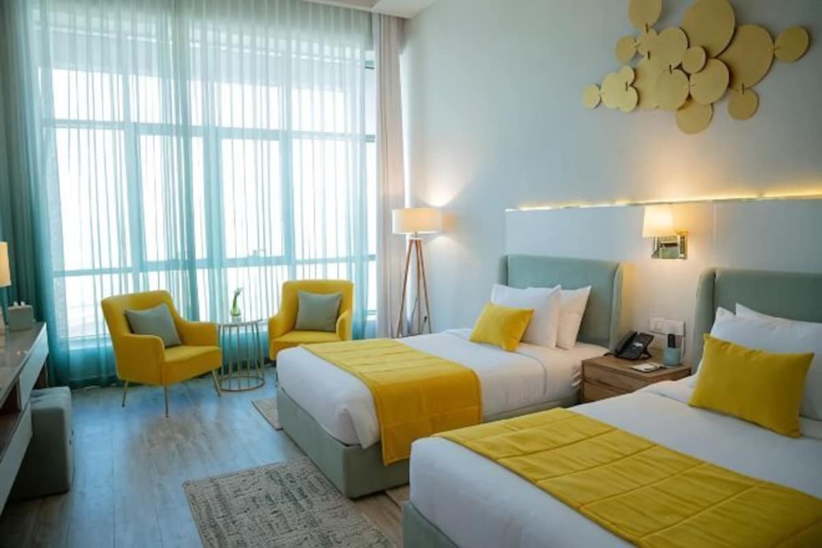 Habitación de hotel con dos camas, un escritorio, una silla, una lámpara y un ventanal grande con cortinas.