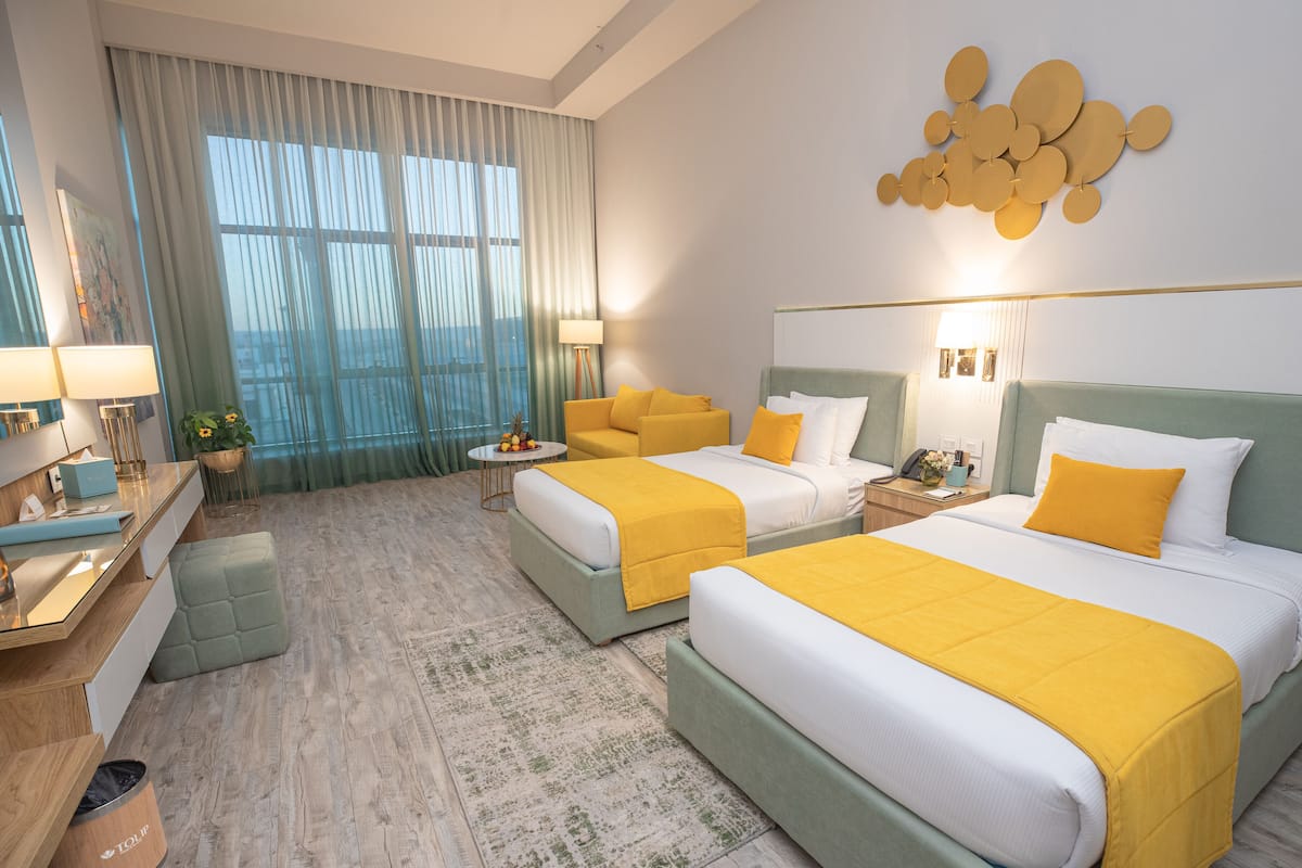 Une chambre d’hôtel avec deux lits, un bureau, une chaise, une petite table avec des fruits et une grande fenêtre avec des rideaux.
