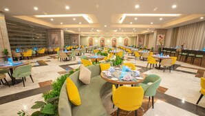 A spacious dining area with multiple tables and chairs, a central seating area with a sofa and armchairs, and a decorative plant arrangement. - Tolip Hotel Olympic International City (New Cairo)