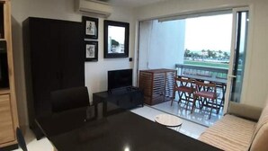 A living room with a flat-screen TV, a wooden cabinet, and a balcony with a table and chairs. - VUE MER Studio Soigné Clim 5 min Plage (Le Lavandou)