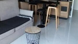 A modern living room with a sofa, a coffee table, and a kitchen area with wooden cabinets and a black countertop. - VUE MER Studio Soigné Clim 5 min Plage (Le Lavandou)