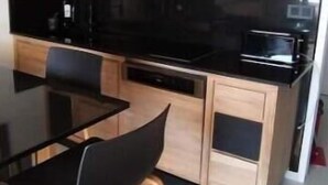 A modern kitchen with wooden cabinets, a black countertop, and a dining table with chairs. - VUE MER Studio Soigné Clim 5 min Plage (Le Lavandou)