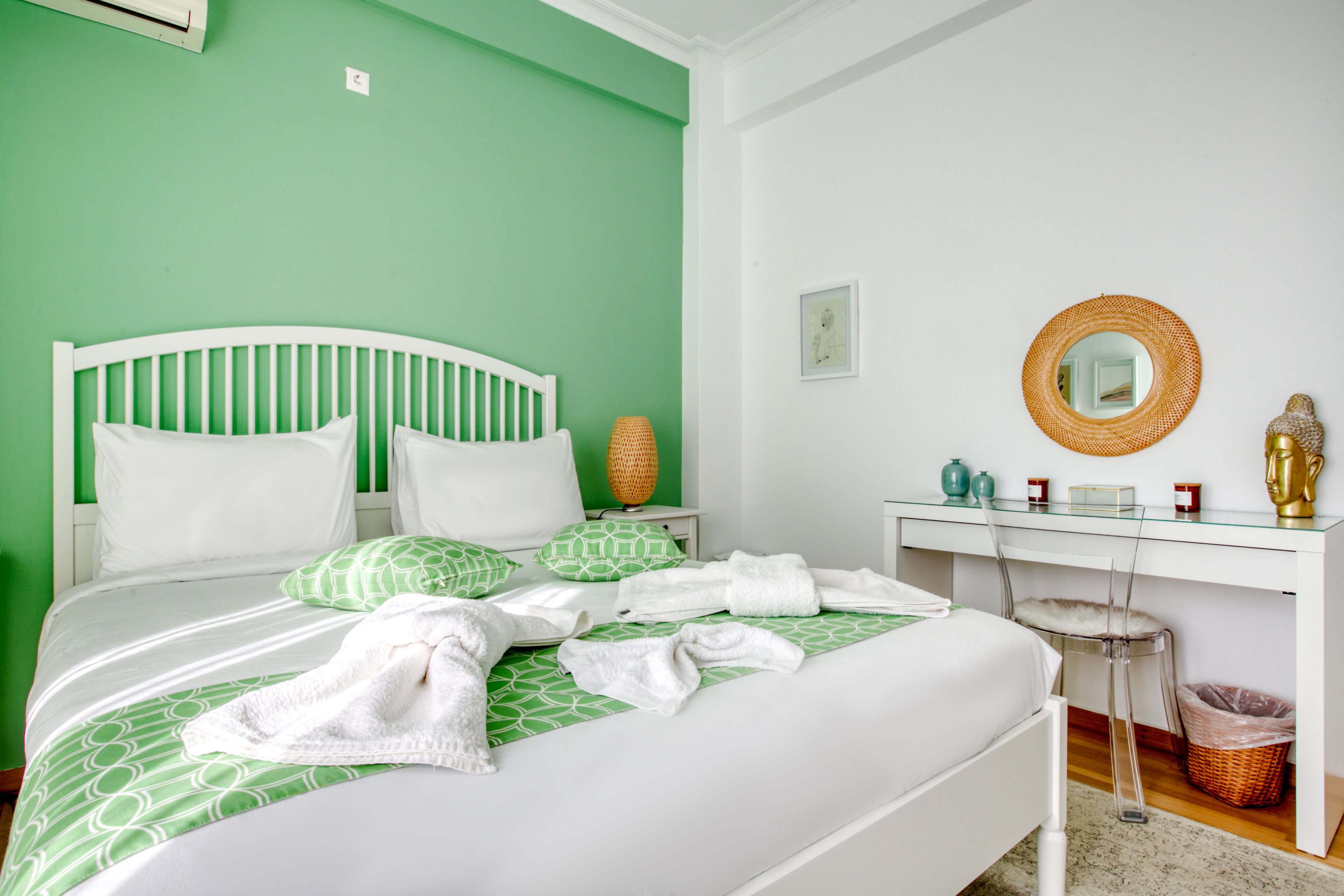 A neatly made bed with green patterned bedding and white pillows, a white bedside table with a lamp, a small framed picture, and a decorative mirror on the wall.