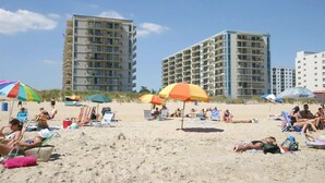 Ein Strand mit mehreren Hochhäusern im Hintergrund, Strandkörben und entspannten Personen im Sand.