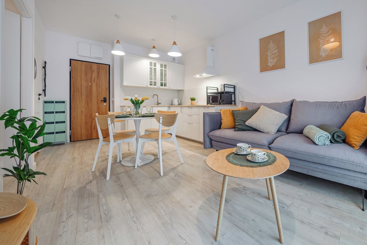 Una sala moderna con un sofá gris, una mesa de comedor de madera y sillas. Se observa un área de cocina con armarios blancos y un microondas.