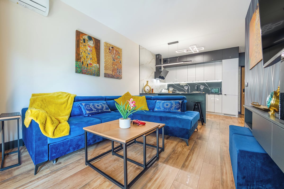 Una sala moderna con un sofá azul, una mesita de madera y una cocina al fondo.