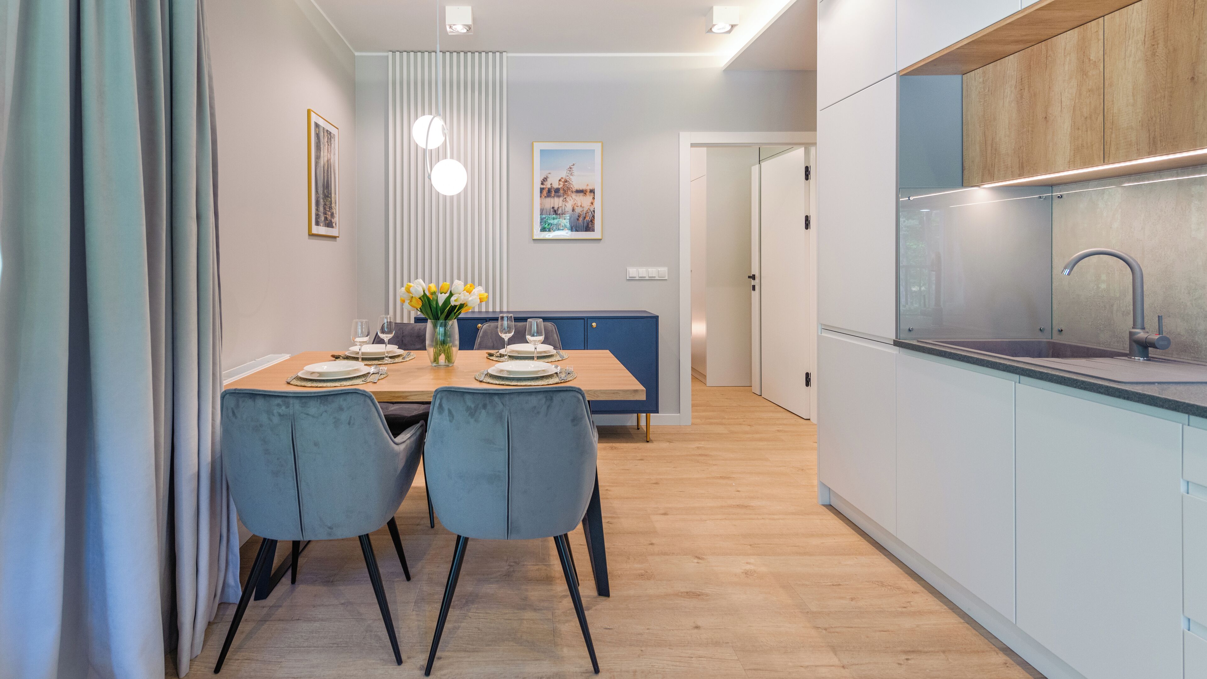 A modern kitchen with a built-in sink, a dining area with a wooden table and blue chairs, and a hallway leading to a door.