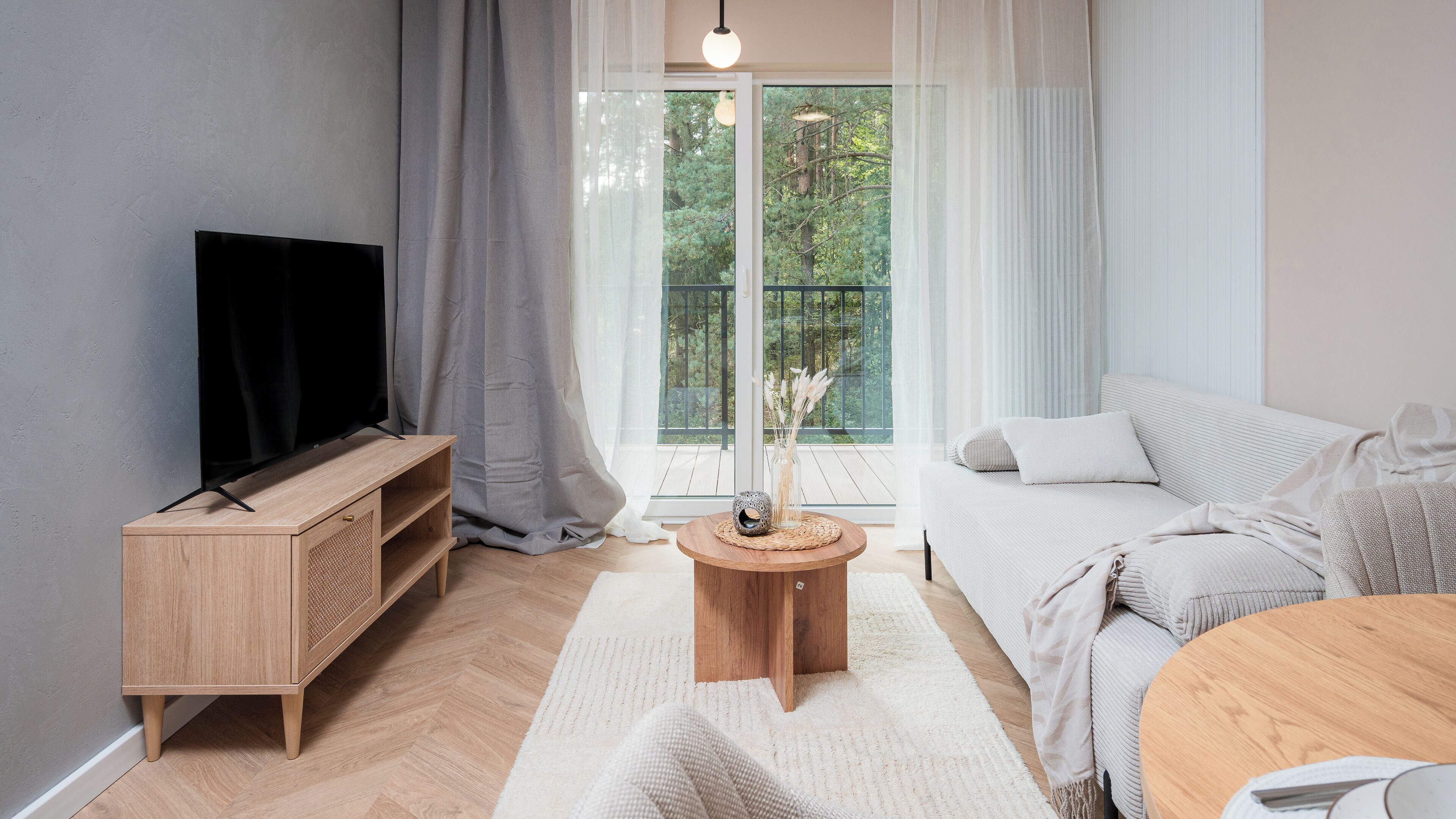 Uma sala de estar moderna com uma TV sobre um suporte de madeira, um sofá, uma mesa de centro redonda de madeira e vista para vegetação através de grandes janelas.