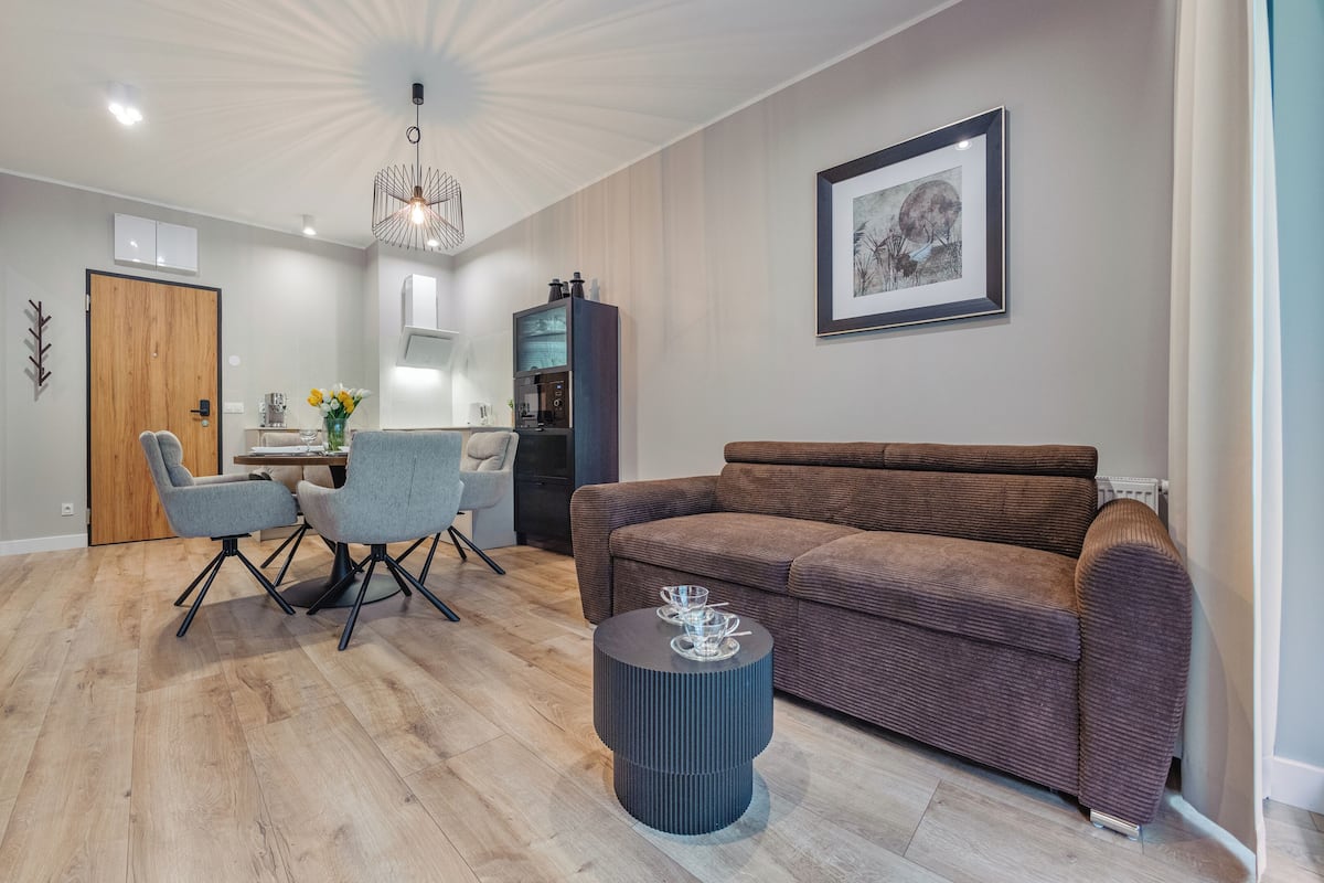 Una sala moderna con un sofá marrón, sillas grises, una mesa de centro, una zona de comedor con mesa y sillas, una cocina y un cuadro en la pared.
