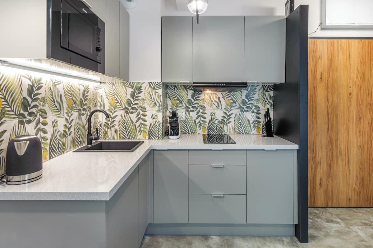 Una cocina con un lavamanos, un hervidor y un microondas. Hay una puerta de madera y una pared con diseño de hojas.