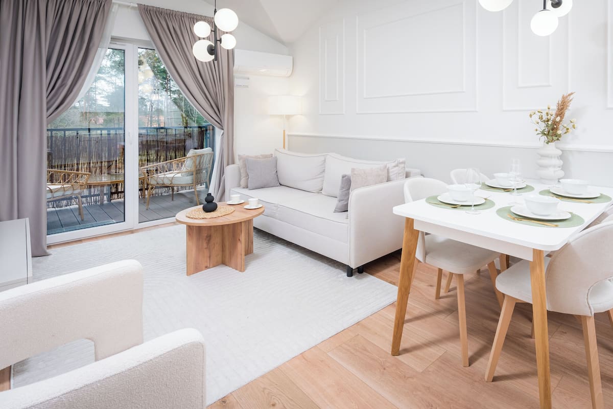 Una sala de estar moderna con un sofá, una mesa de comedor preparada para cuatro personas y un ventanal con cortinas.