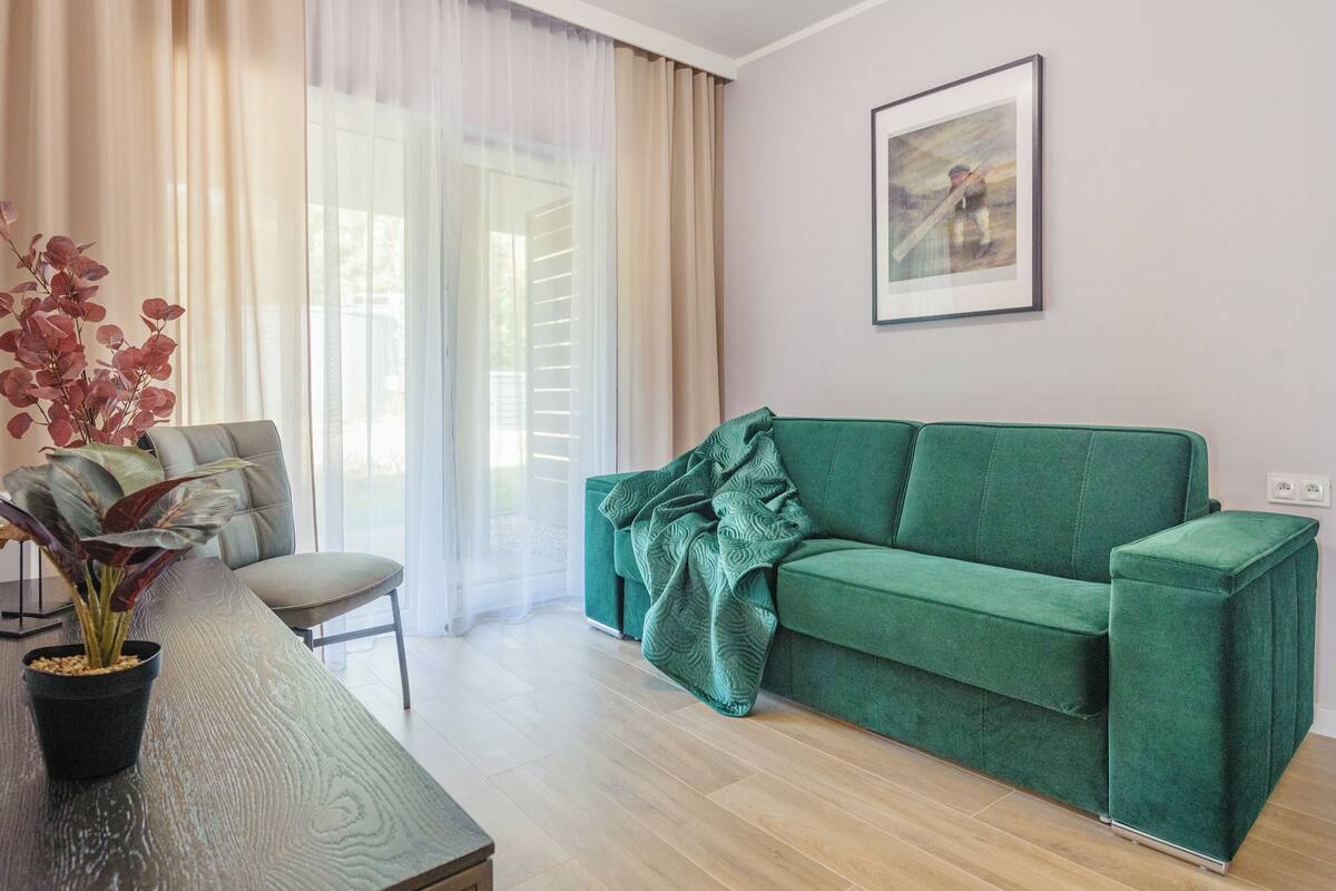 Una sala de estar con un sofá verde, una mesa de comedor con una planta en maceta y una ventana con cortinas ligeras.