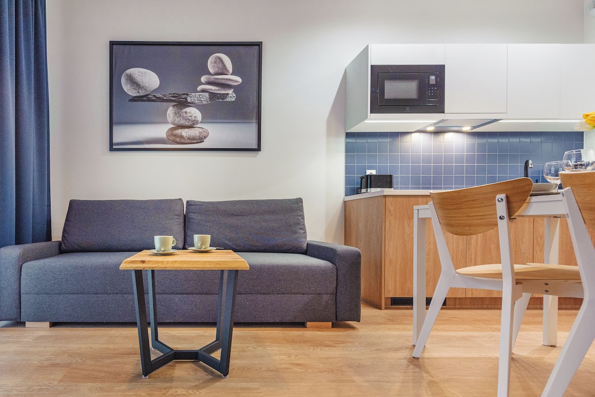 Un espacio moderno con sala de estar, un sofá, una mesita con tazas, una cocina con microondas y un salpicadero de azulejos azules.
