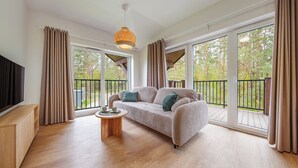 Ein geräumiges Wohnzimmer mit einer Couch, einem Couchtisch und einem Fernseher. Gro ße Fenster bieten Blick auf Bäume und einen Balkon.