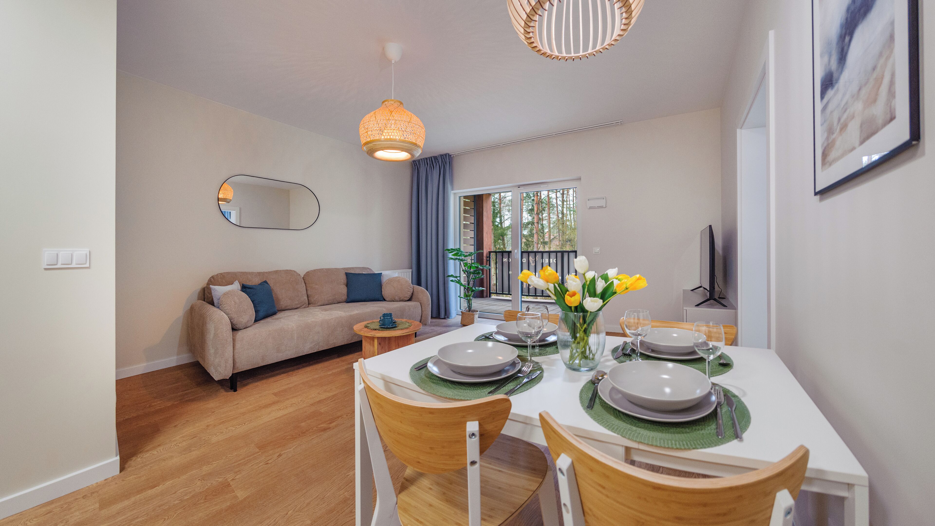 A dining area with a table set for two, a sofa, a television, and a balcony with a view.