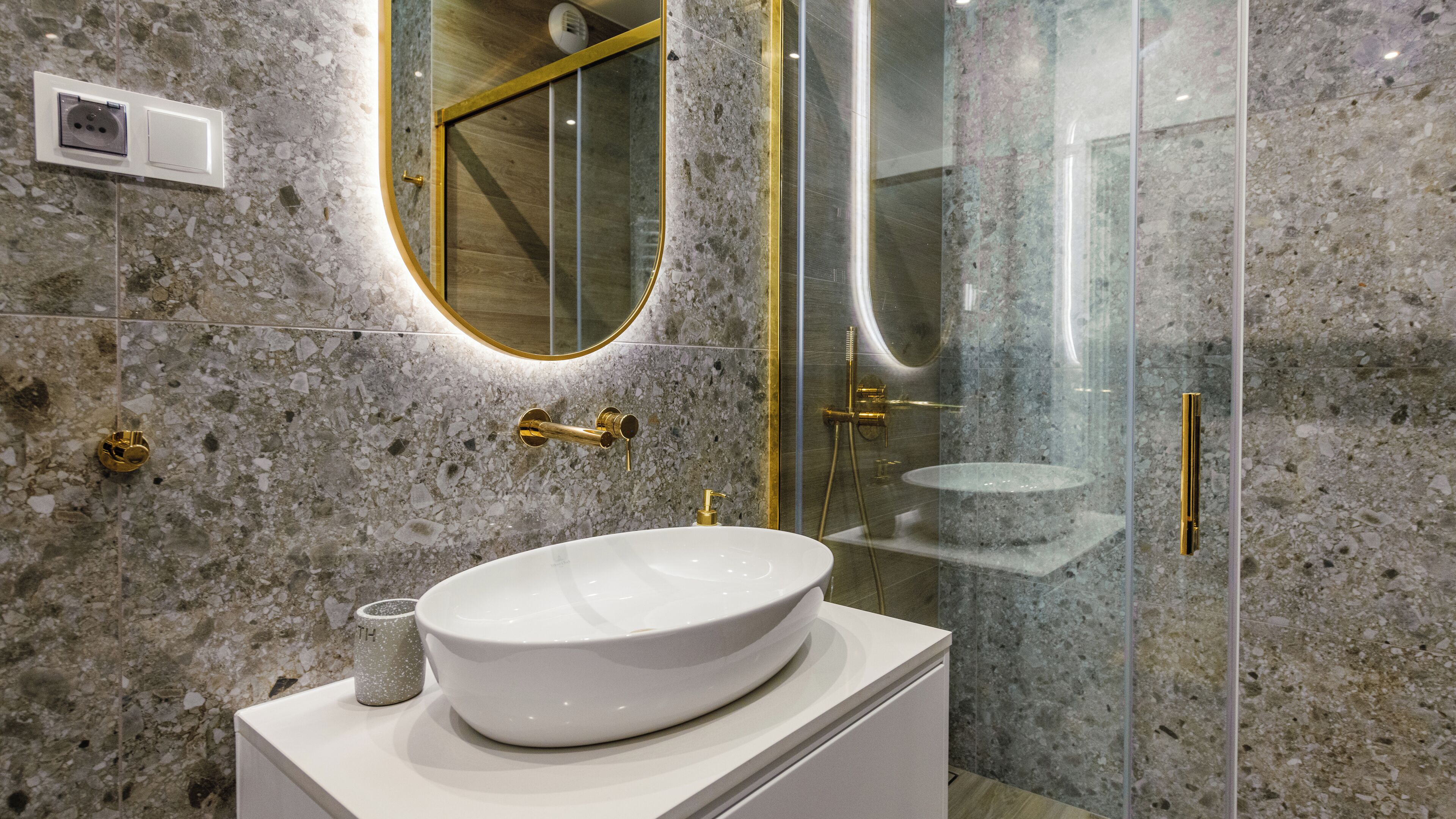 Un baño con lavamanos blanco, detalles dorados, cabina de ducha con mampara de vidrio y pared de mármol.