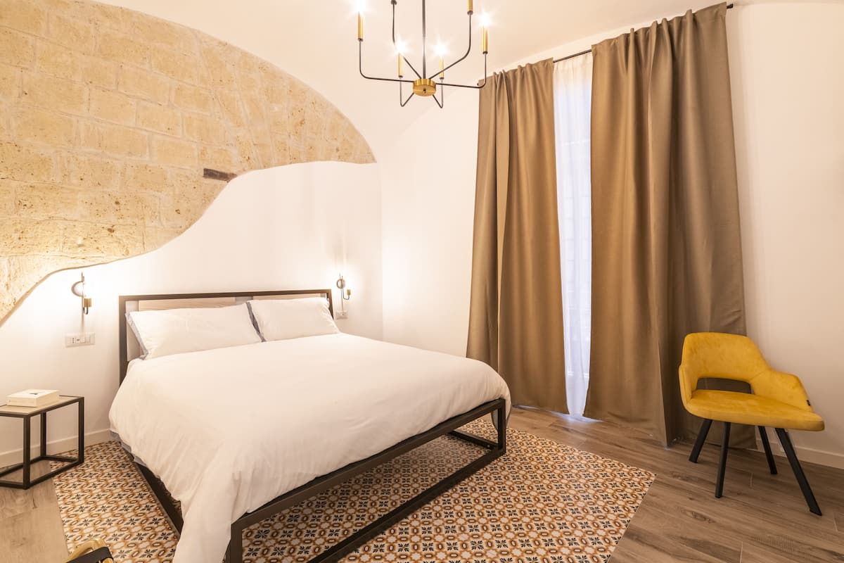 Un dormitorio con cama, una silla amarilla, alfombra estampada, pared de ladrillo y un candelabro.
