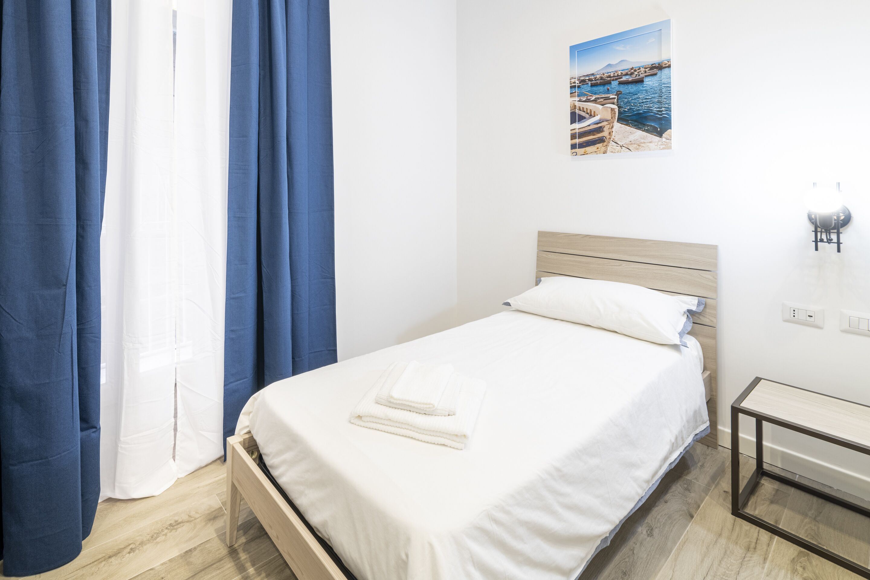 Quarto com cama de madeira, roupa de cama branca, mesa de cabeceira pequena e um quadro na parede.