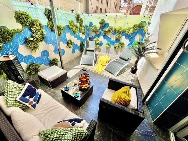 Un espace extérieur ensoleillé, aménagé avec des sièges, un mur recouvert de carreaux bleus et blancs, une petite table sur laquelle sont disposés des fruits et des fauteuils confortables.