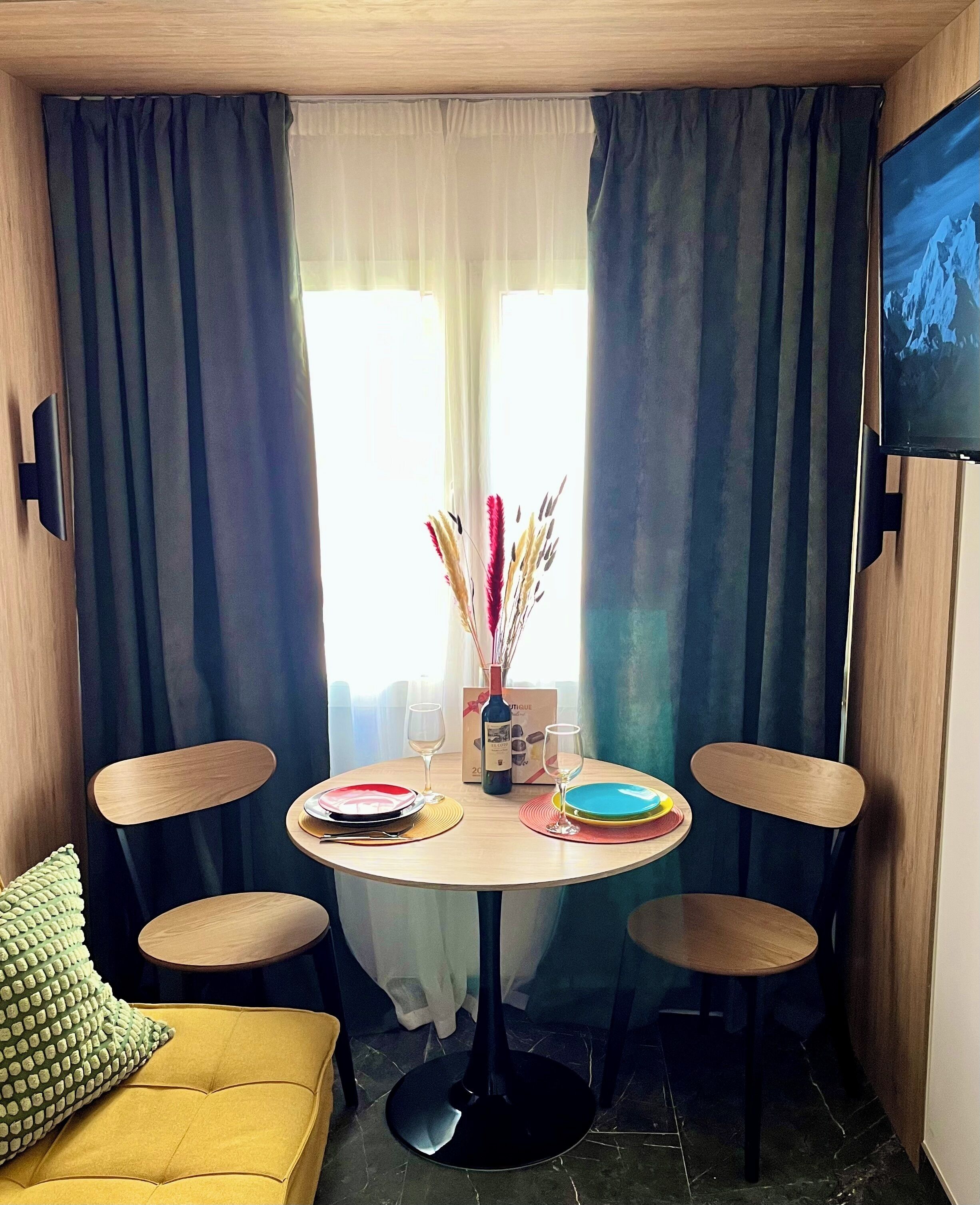 Ein gemütlicher Raum mit rundem Tisch für zwei Personen, einer Couch und einem Fernseher an der Wand.