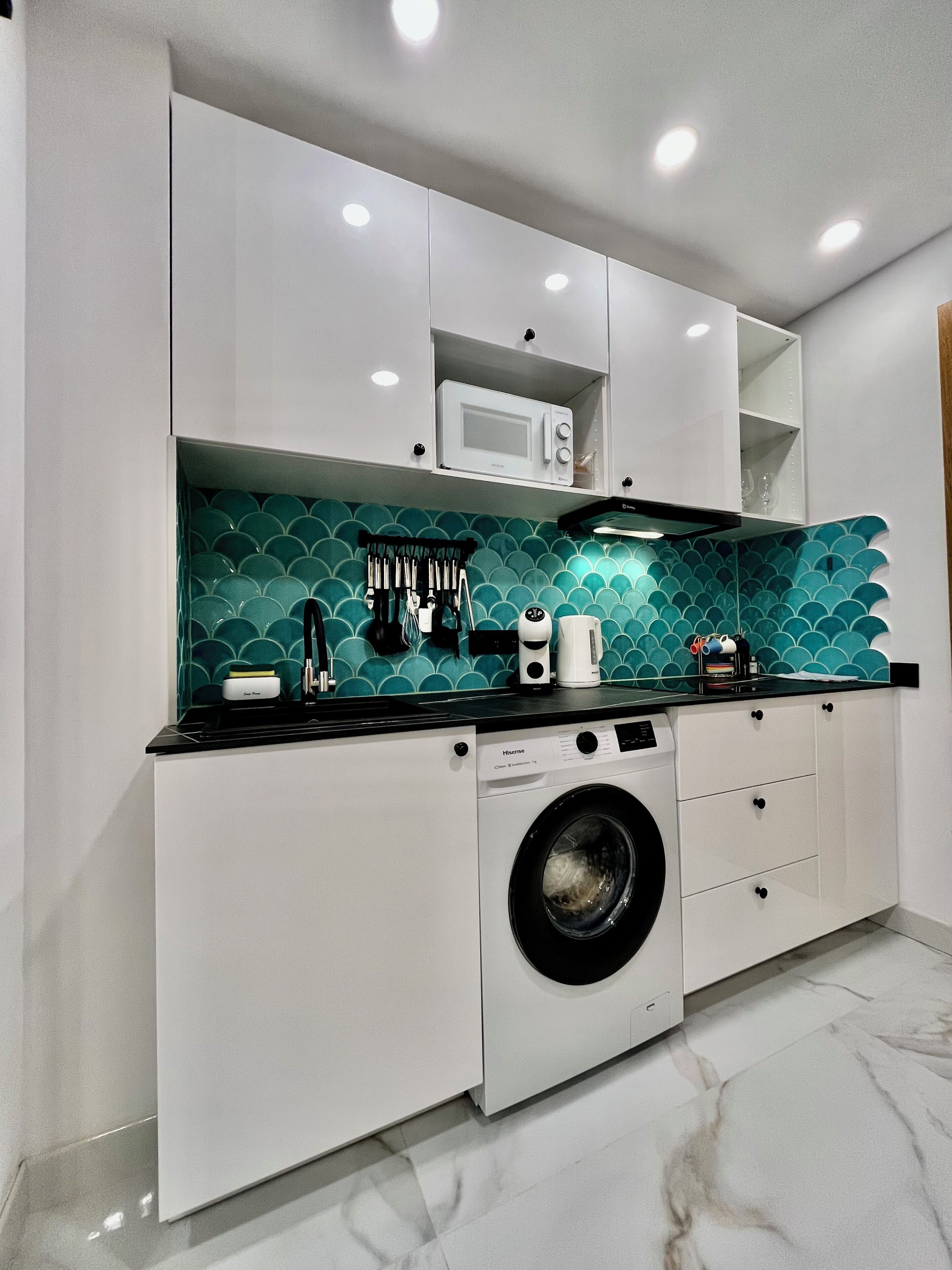 Een compacte keuken met een wasmachine, magnetron en een blauwgroene spatwand.