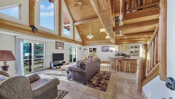 A spacious living area with a high ceiling, wooden beams, a sofa, a television, and a kitchen area with stools.