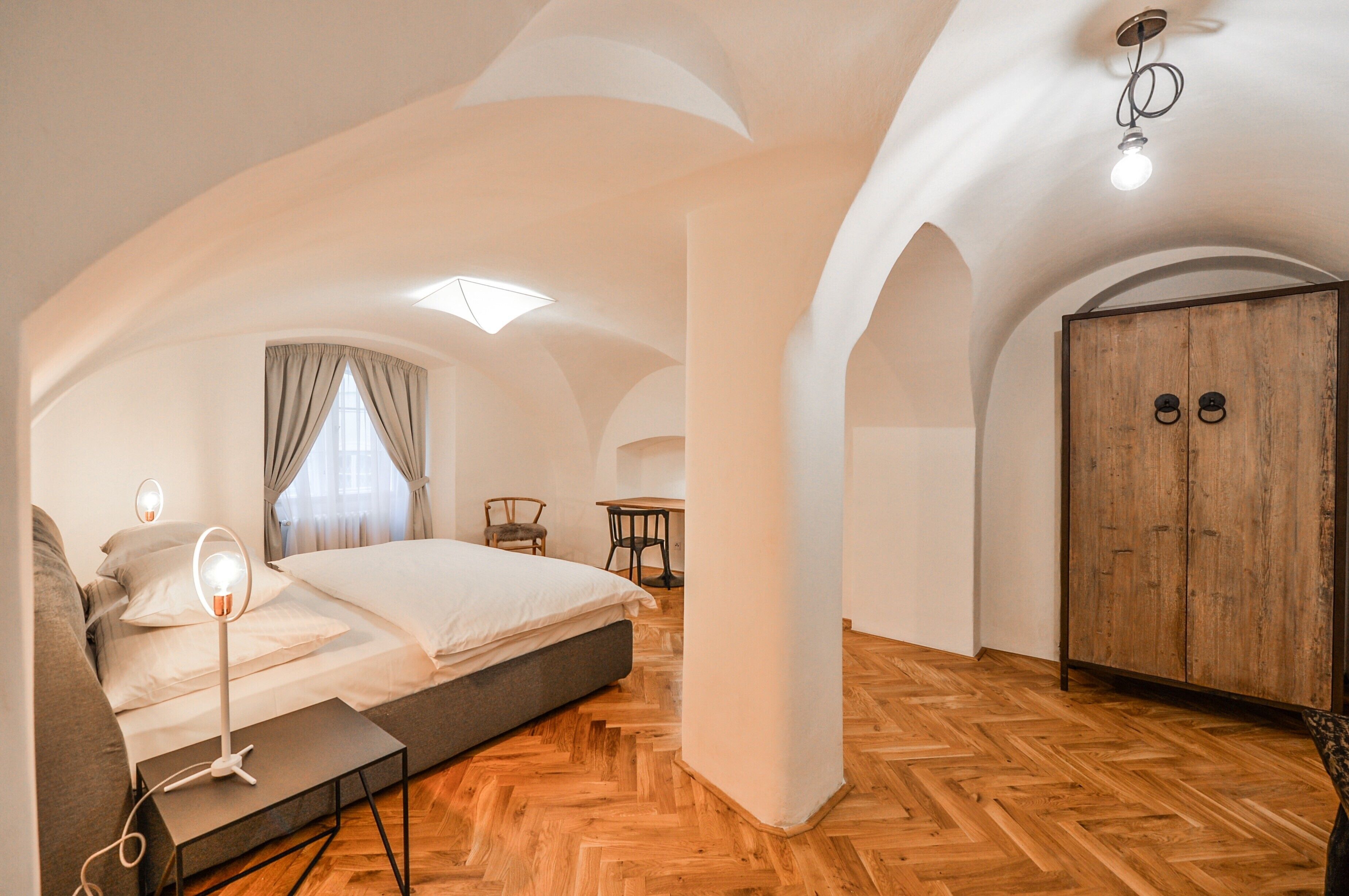 Un dormitorio con cama, mesita de noche, lámpara, armario de madera y ventana con cortinas.