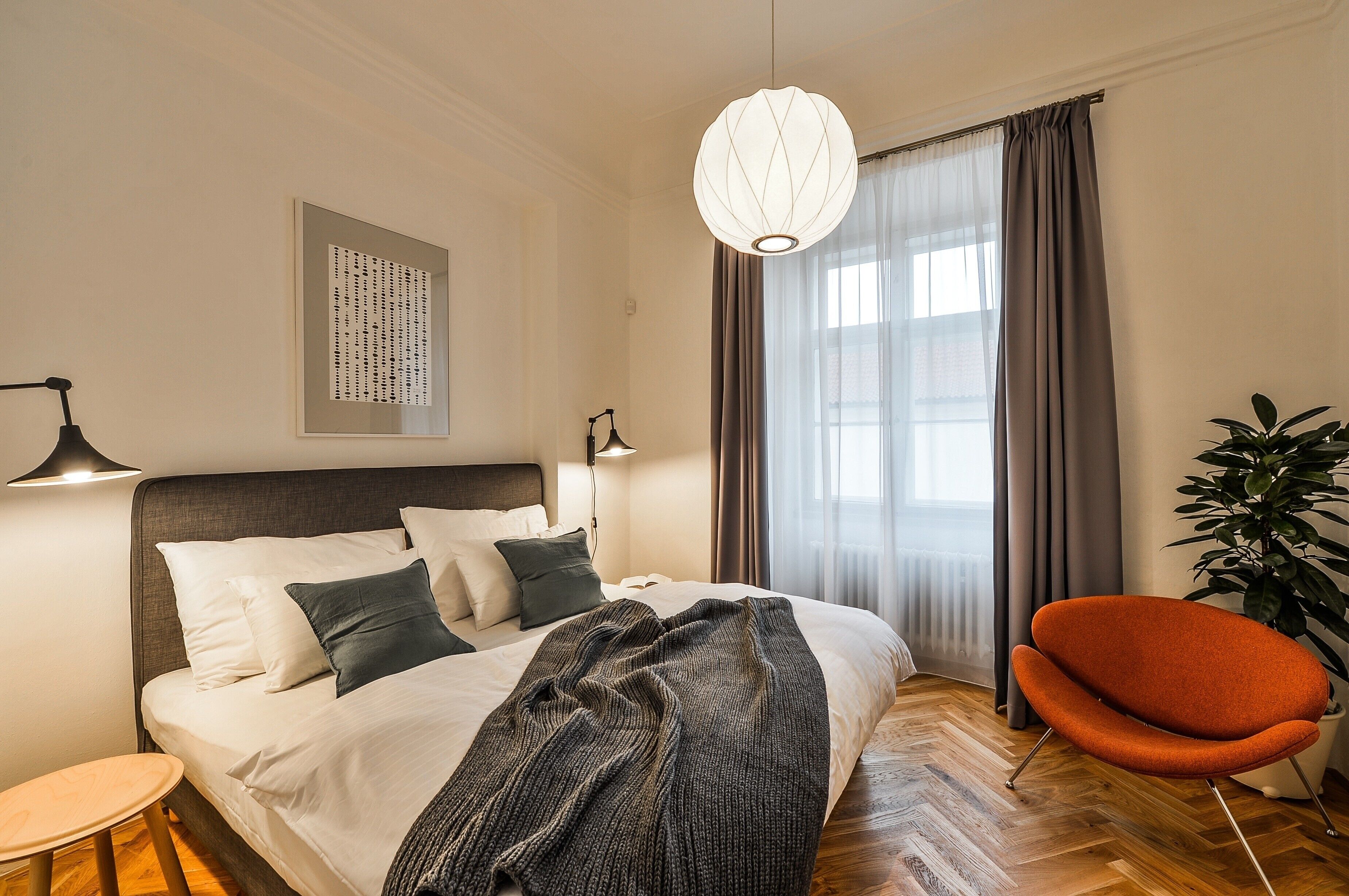 A modern hotel room with a large bed, a bedside table, a reading lamp, a potted plant, and a view of the outdoors through a window with curtains.