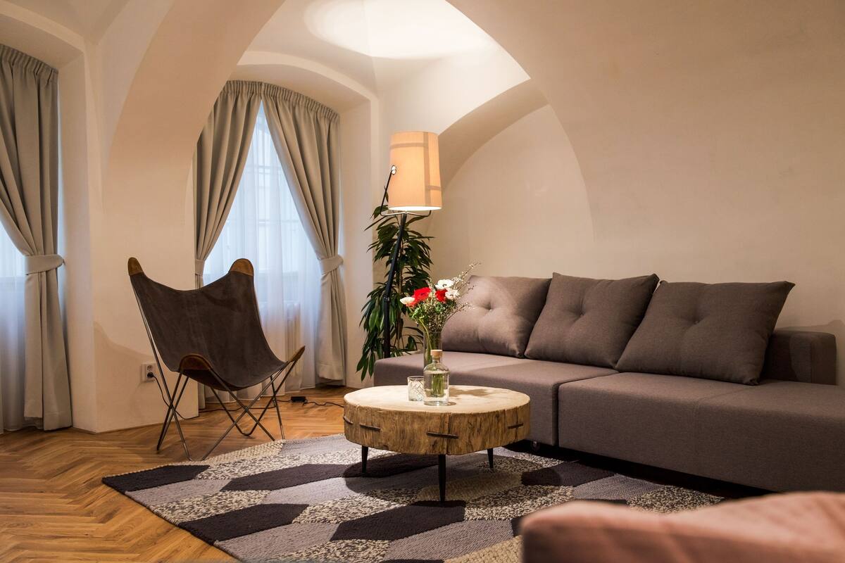 A living room with a round wooden coffee table, a grey sofa, a brown leather chair, a floor lamp, and a vase with flowers.
