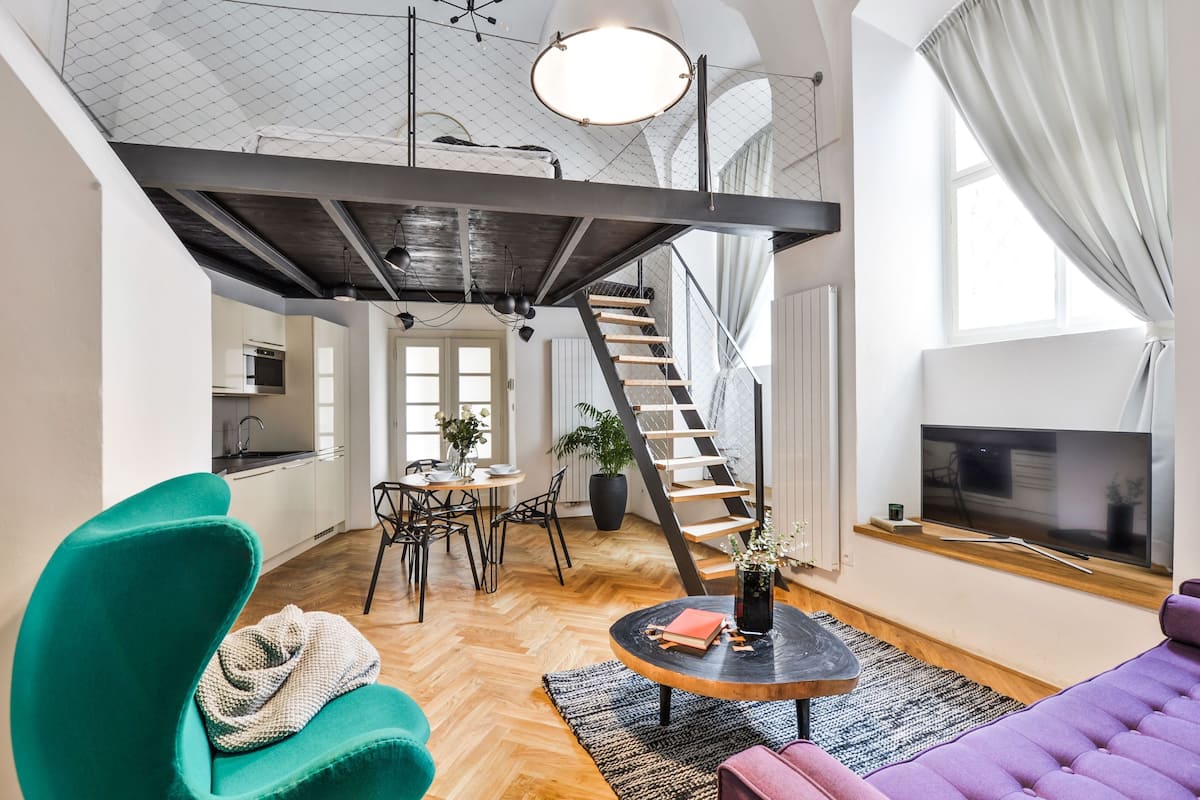 A spacious loft apartment with a high ceiling, a staircase, a dining area, and a living room with a large TV.
