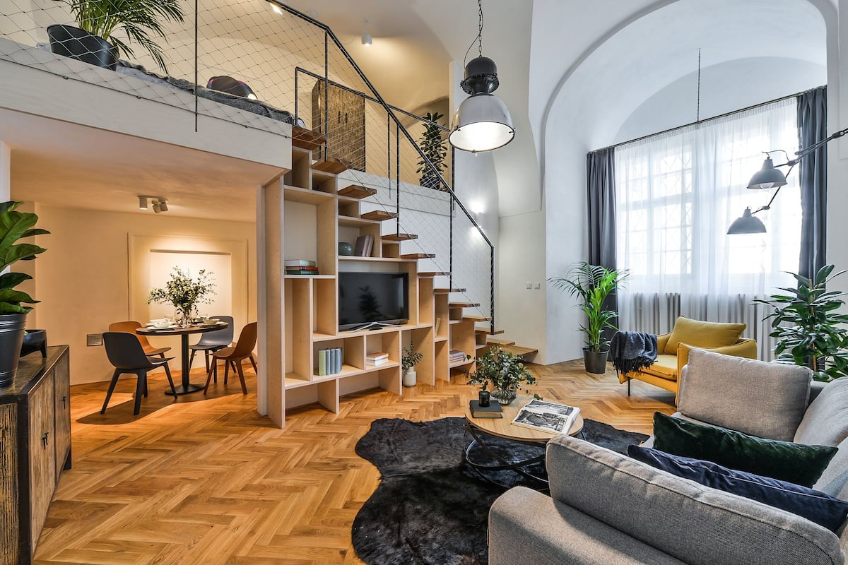 A spacious living area with a herringbone wood floor, a large sofa, a dining table with chairs, a bookshelf, and a TV.
