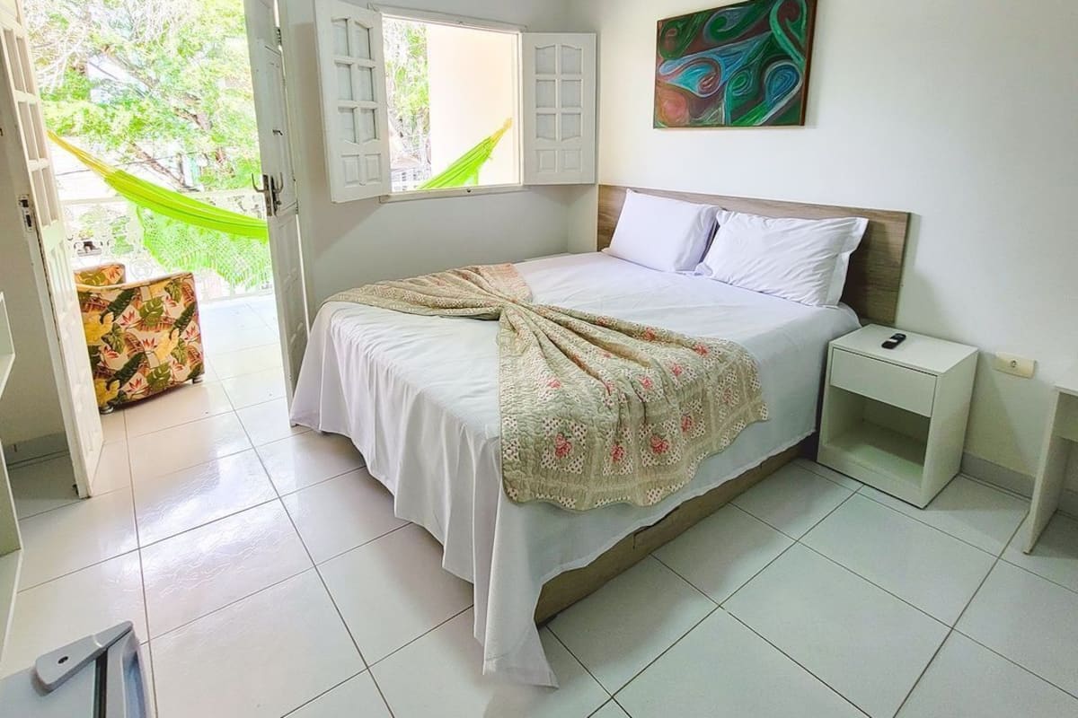 A bedroom with a bed, a nightstand, a window with open shutters, a hammock outside, and a painting on the wall.