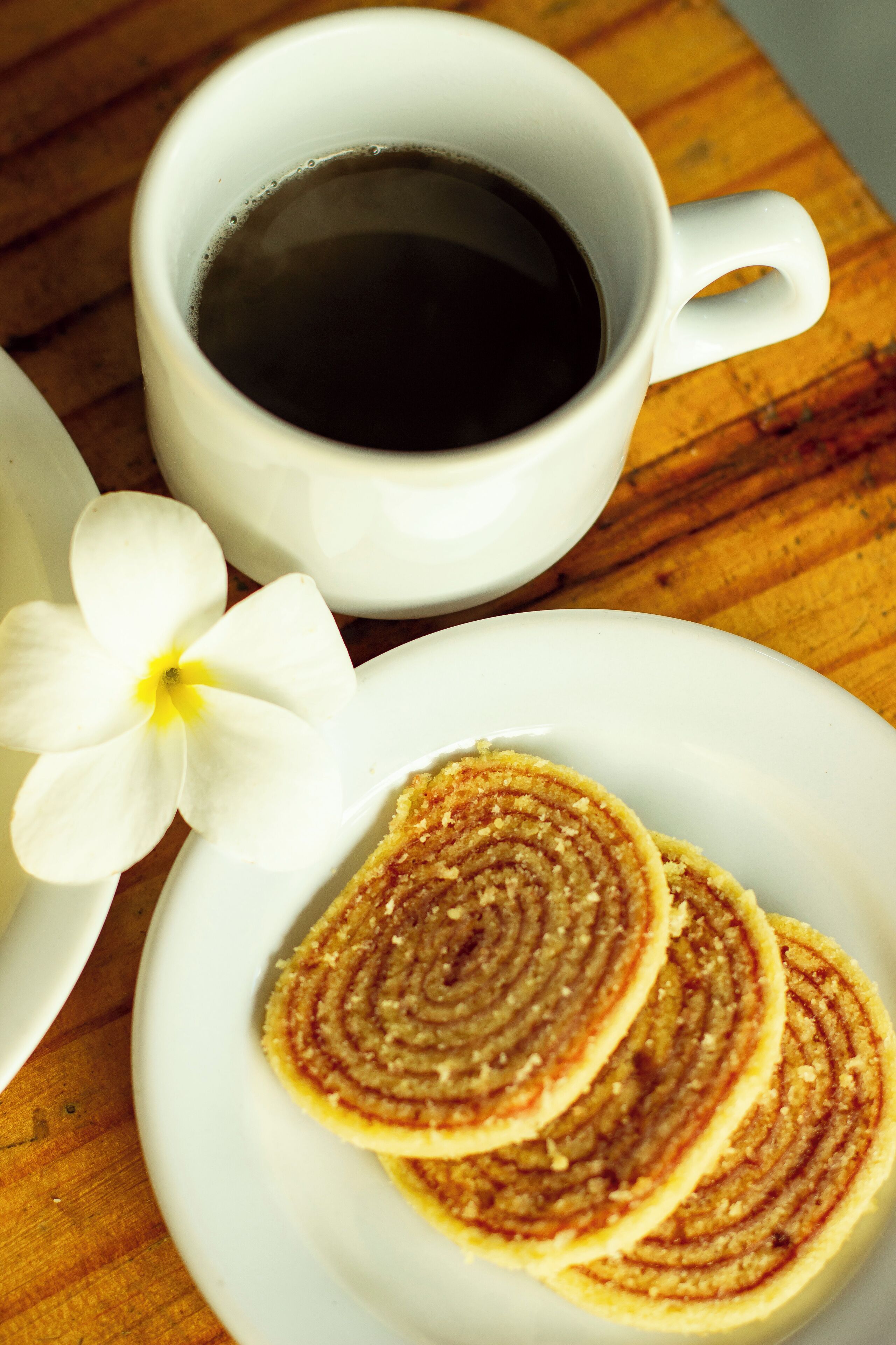 Une tasse de café blanche, une fleur blanche et une assiette contenant trois pâtisseries en forme de spirale, le tout posé sur une surface en bois.
