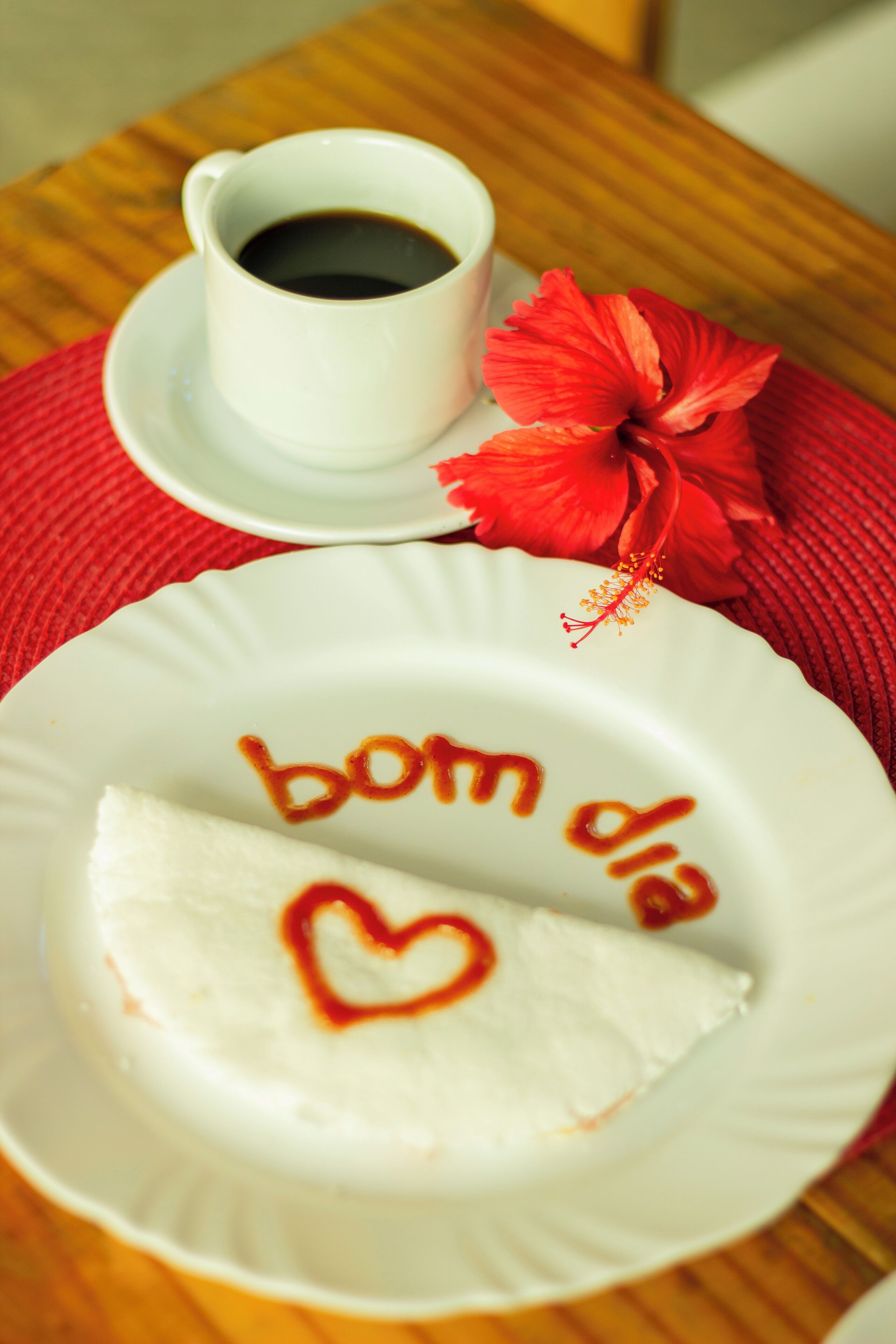 Un plato con diseño de corazón y la inscripción 'bom dia', una taza de café y una flor roja sobre una mesa.