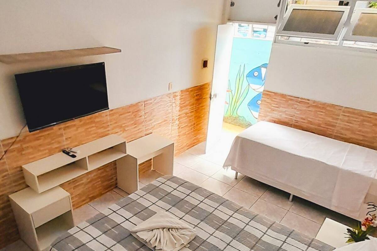 A bedroom with a bed, a TV mounted on the wall, a bench, and a window with a view of a blue sky and greenery.