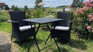 A garden patio with two wicker chairs and a table, surrounded by lush greenery and flowering plants.
