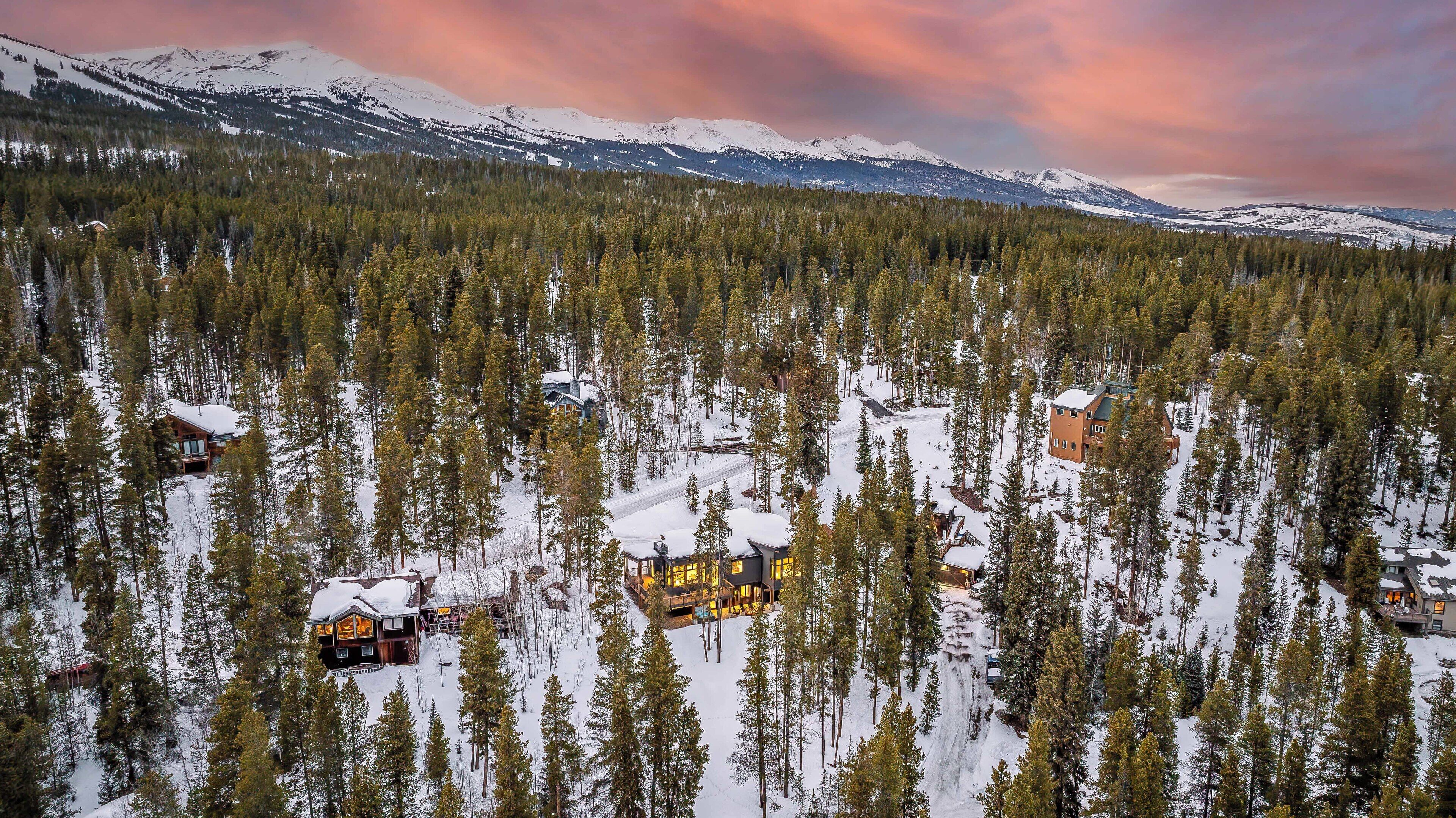 A snow-covered forest with several cabins and a lodge, surrounded by mountains and a sunset sky.