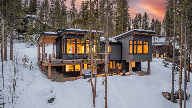 Une cabane moderne à plusieurs niveaux, dotée de grandes fenêtres et d’une terrasse, entourée de neige et d’arbres.