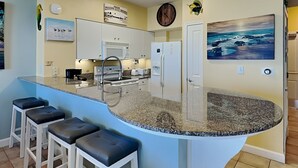 A kitchen with a curved island, white bar stools, a microwave, a refrigerator, and a painting of the ocean. - Shores of Panama #1129 | Stunning Views + Fun On-Site Amenities! (Panama City Beach)