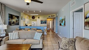 A spacious living area with a sofa, a coffee table, and a rug. A kitchen is visible in the background with a sign that reads "Beach." - Shores of Panama #1129 | Stunning Views + Fun On-Site Amenities! (Panama City Beach)