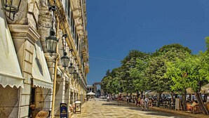 A historic street with stone buildings, outdoor seating, and trees.