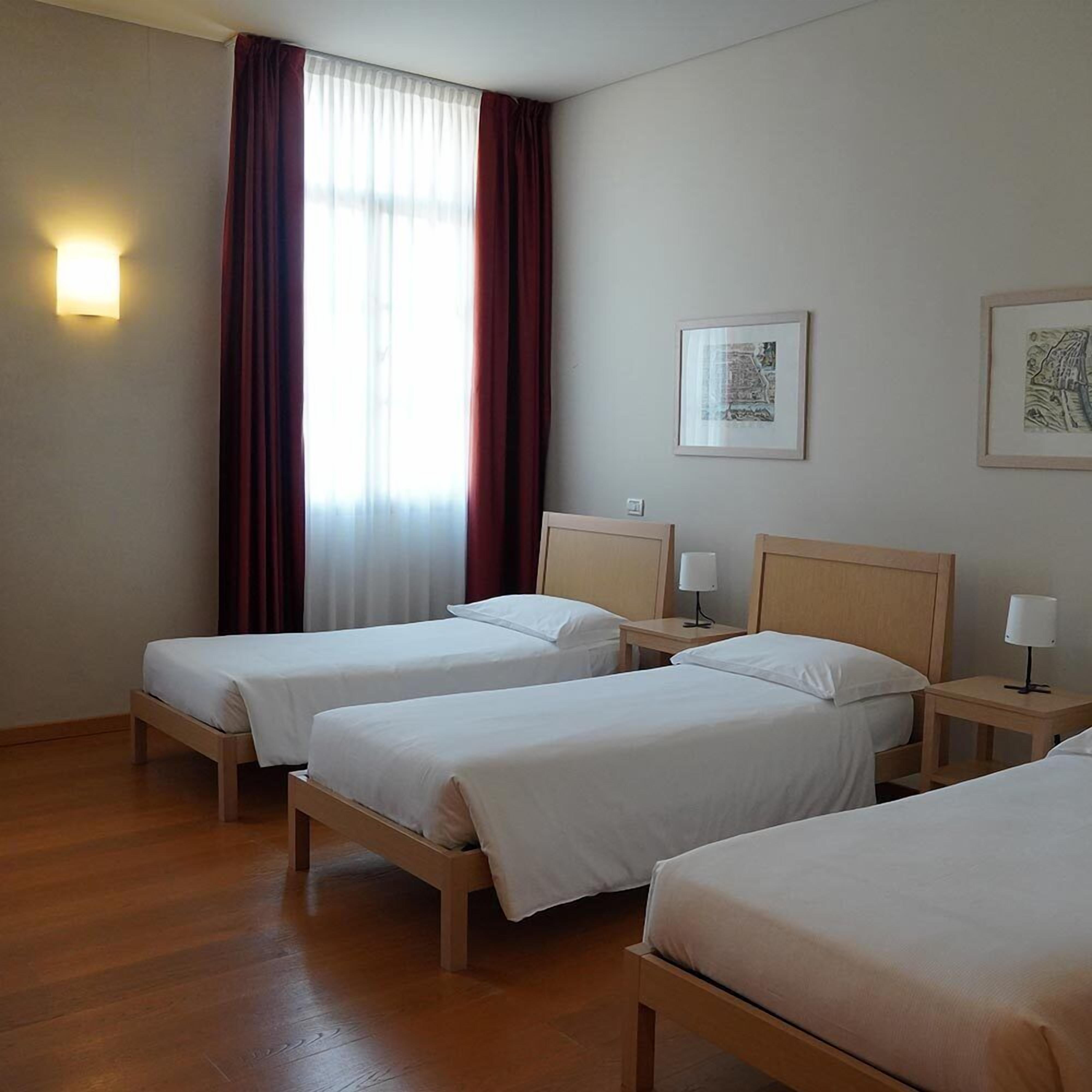 Quarto de hotel com duas camas de solteiro, piso de madeira, janela com cortinas, luminária fixada na parede e dois quadros emoldurados.