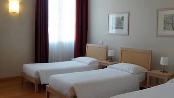 Quarto de hotel com duas camas de solteiro, piso de madeira, janela com cortinas, luminária fixada na parede e dois quadros emoldurados.