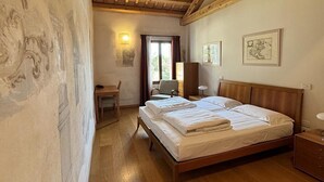 A bedroom with a wooden ceiling, a bed with white linens, a desk with a chair, and two framed artworks on the wall.