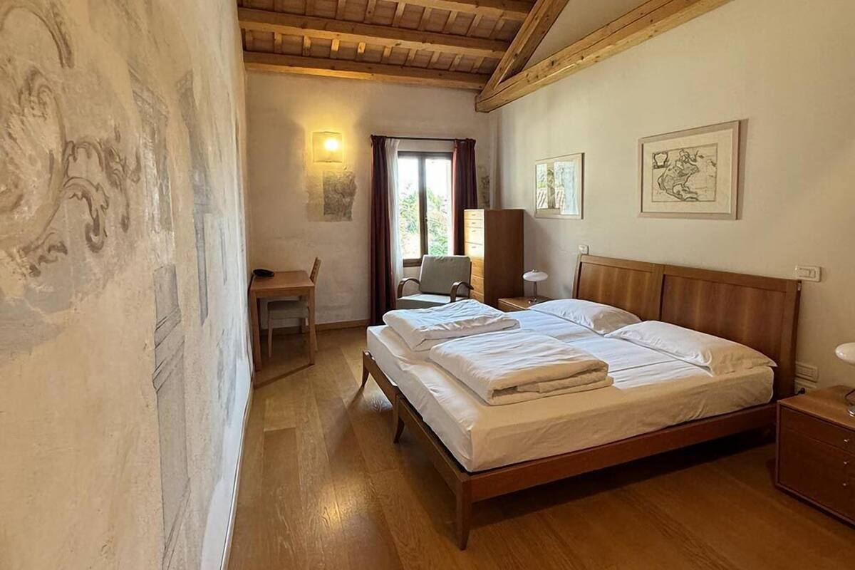 A bedroom with a wooden ceiling, a bed with white linens, a desk with a chair, and two framed artworks on the wall.